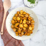 air fryer breakfast potatoes on white serving plate next to wooden serving spoon. Dish of sliced chives in the top right corner.