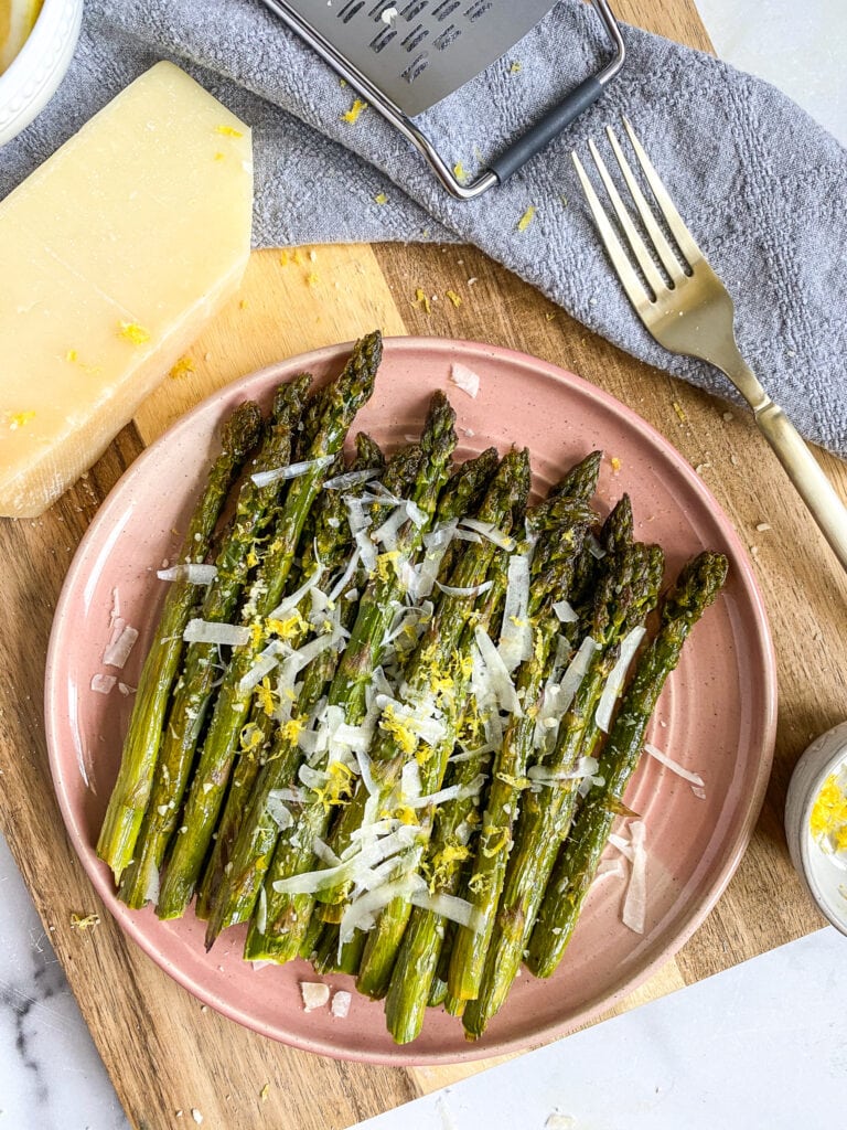 lemon parmesan asparagus on a pink ceramic plate resting on a wooden cutting board with a fork in the background & a wedge of Parmesan Cheese