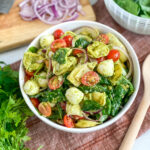 pesto tortellini pasta salad in white serving bowl with parsley in the background. There is also a wooden cutting board with sliced red onion and a chefs knife in the background