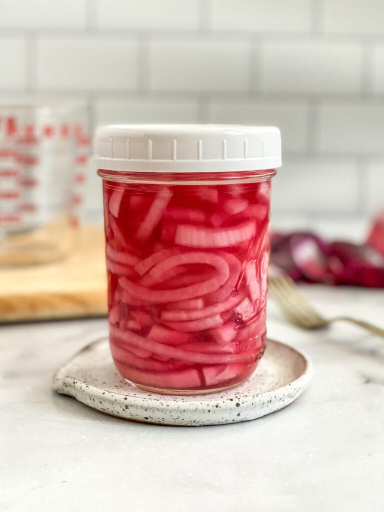 quick pickled onions in a mason jar with a white plastic lid. Jar is sitting on a spoon rest & there are onion skins and a fork in the background.