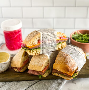roast beef sandwiches, sliced and wrapped in newspaper. Sandwiches are stacked up on a cutting board with leftover ingredients in the background.