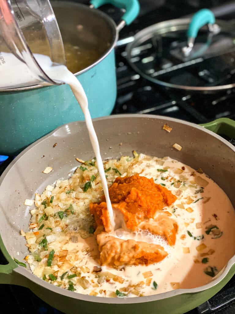 Saute pan with onions, garlic and sage cooked together. Pumpkin puree is on top and milk is being slowly added to the pan.