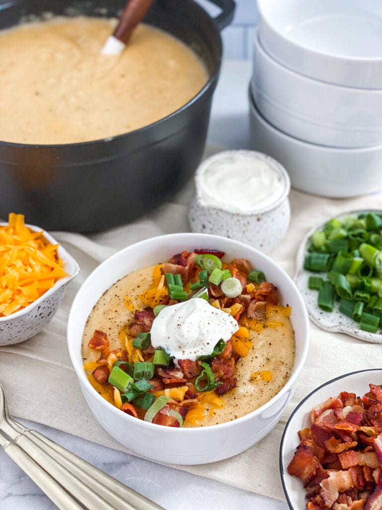 cheesy loaded potato soup served up in a bowl woth cheddar cheese, bacon, scallions and sour cream on top. The extra toppings are surrounding the bowl on small plates and the soup pot is in the background