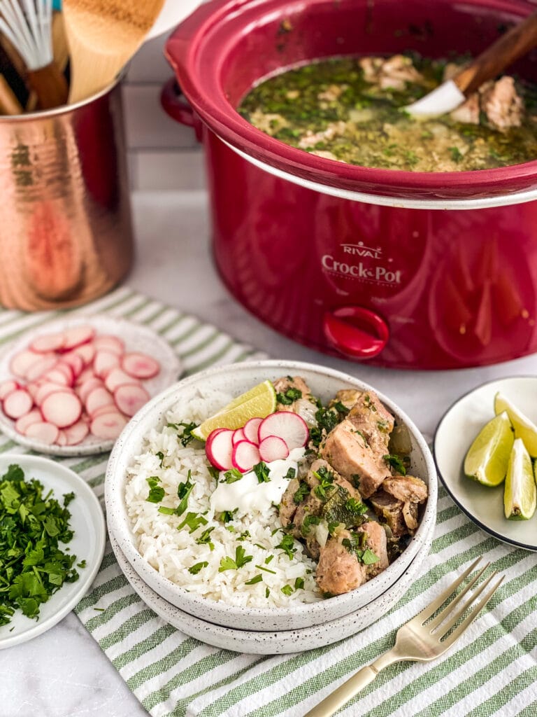 salsa verde pork plated up with rice, cilantro, lime and sour cream. Crockpot in the background with remaining pork and extra ingredients are also in frame surrounding the plate of food.
