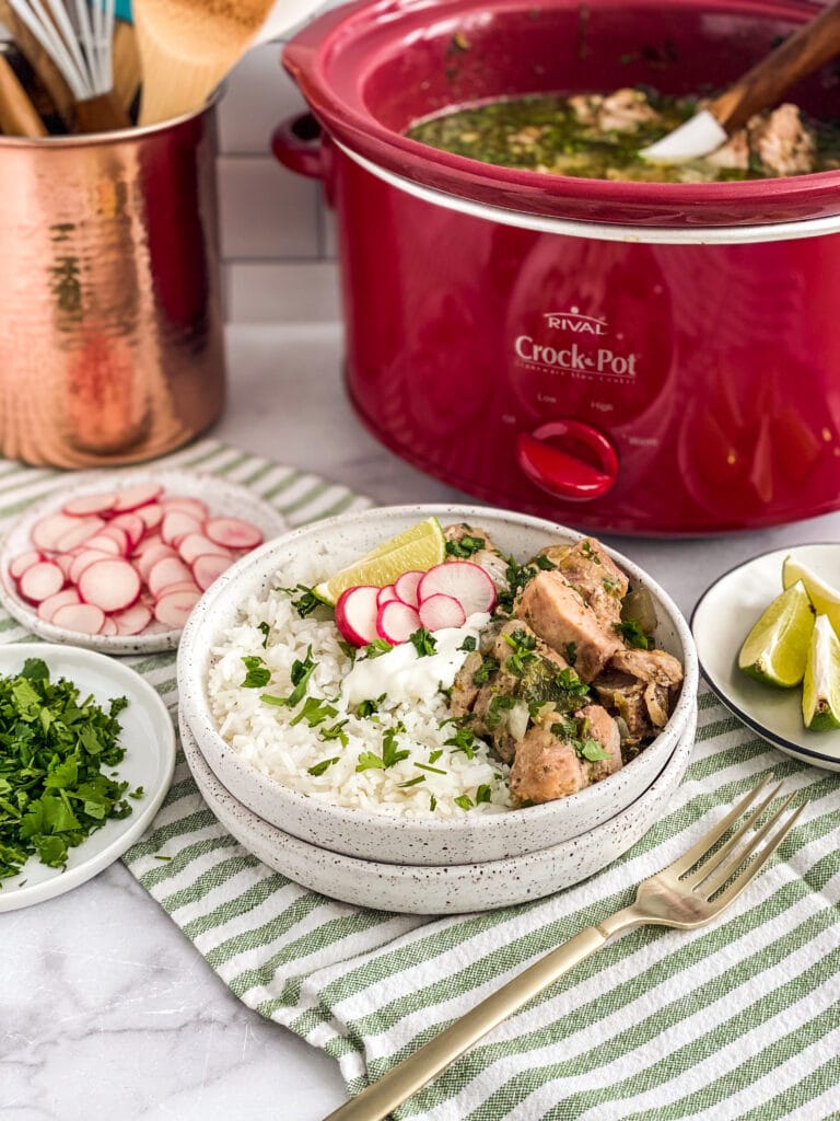 salsa verde pork plated up with rice, cilantro, lime and sour cream. Crockpot in the background with remaining pork and extra ingredients are also in frame surrounding the plate of food.