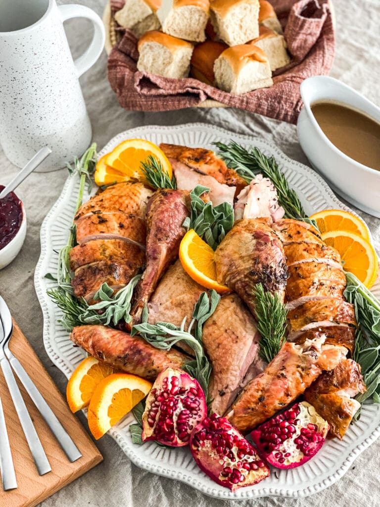 whole roasted turkey, carved on a serving platter with herbs and citrus to garnish the plate. Gravy, cranberry sauce & rolls are in the background of the image.
