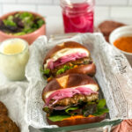 final shot of lamb burger, on a pretzel bun, topped with mixed greens, paprika mayo, whipped feta and pickled onions. burgers are sitting in a glass loaf pan lined with newspaper. Extra ingredients are in the background of the image.