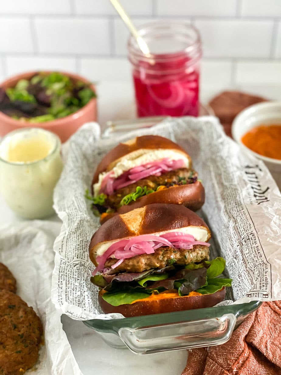 final shot of lamb burger, on a pretzel bun, topped with mixed greens, paprika mayo, whipped feta and pickled onions. burgers are sitting in a glass loaf pan lined with newspaper. Extra ingredients are in the background of the image.